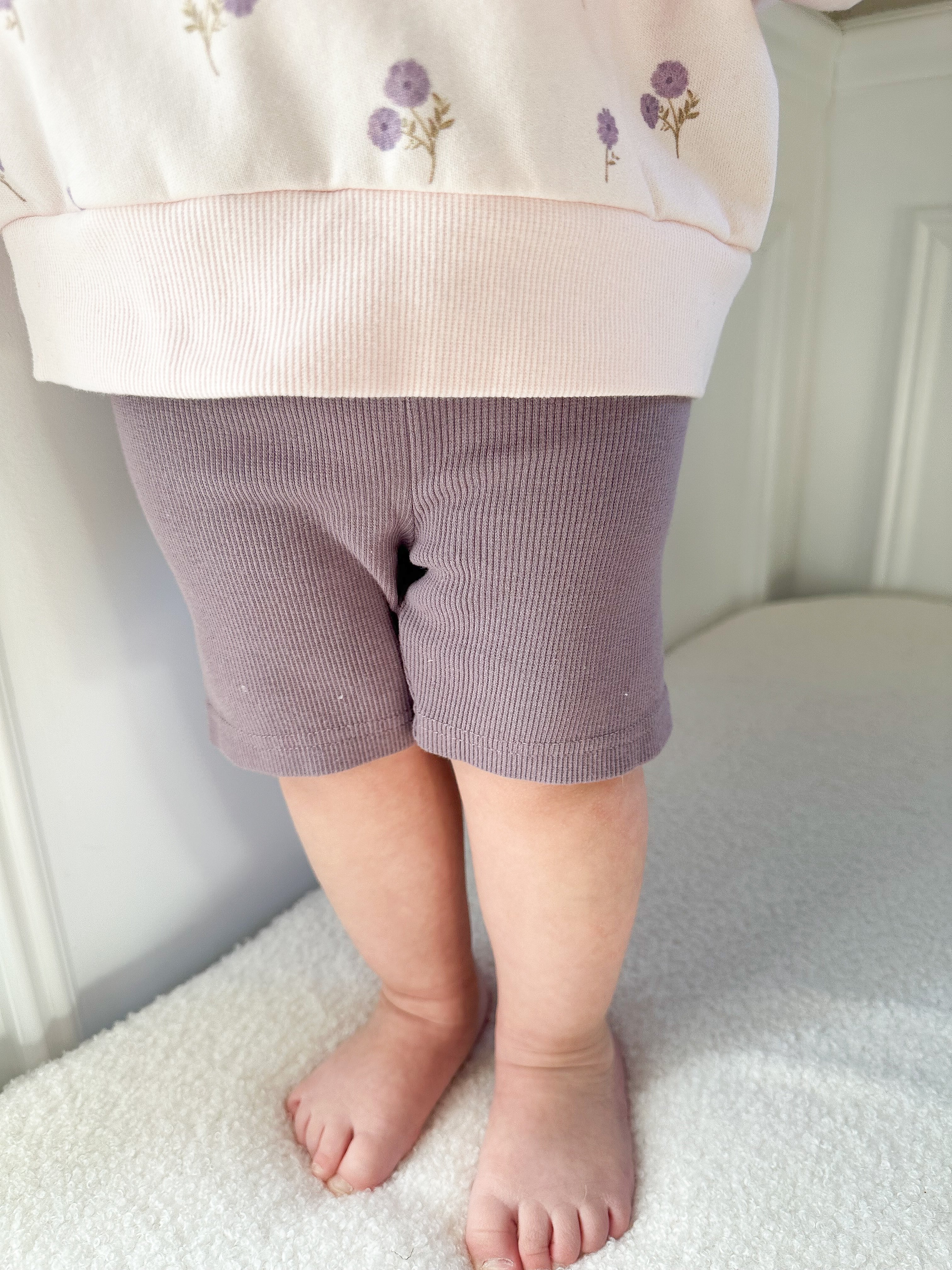 Child wearing purple ribbed biker shorts and light-colored sweatshirt with purple floral pattern standing on beige surface.
