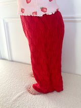 Child wearing dark pink knitted pants and a tank top with strawberry pattern standing on a light surface. Cropped to only show pants and small part of tank top.