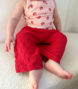Child wearing a pink top with strawberry pattern and dark pink knitted pants sitting on a white surface. Cropped to only show outfit.