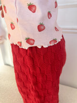 Child's wearing pink tank top with strawberry pattern and red textured pants on a neutral background. Cropped to show bottom of tank top and top of pants.