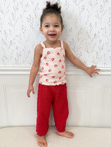 Child wearing a strawberry-patterned tank top and red pants standing against a white molding with subtle floral design above. Child is smiling and gently touching molding.