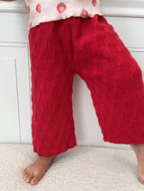 Dark pink knitted pants worn by a child standing on a light colored surface against a white background. 