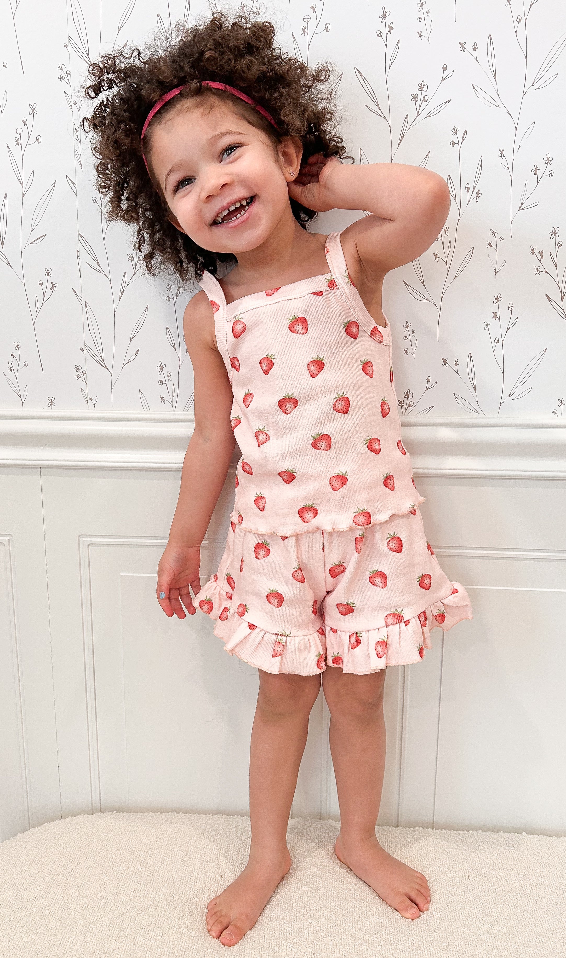Child wearing a strawberry-patterned outfit standing against a white wall with floral decorations.