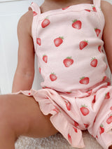 Child sitting on light-colored surface wearing a pink outfit with strawberry pattern on a white background. Cropped to only show outfit.