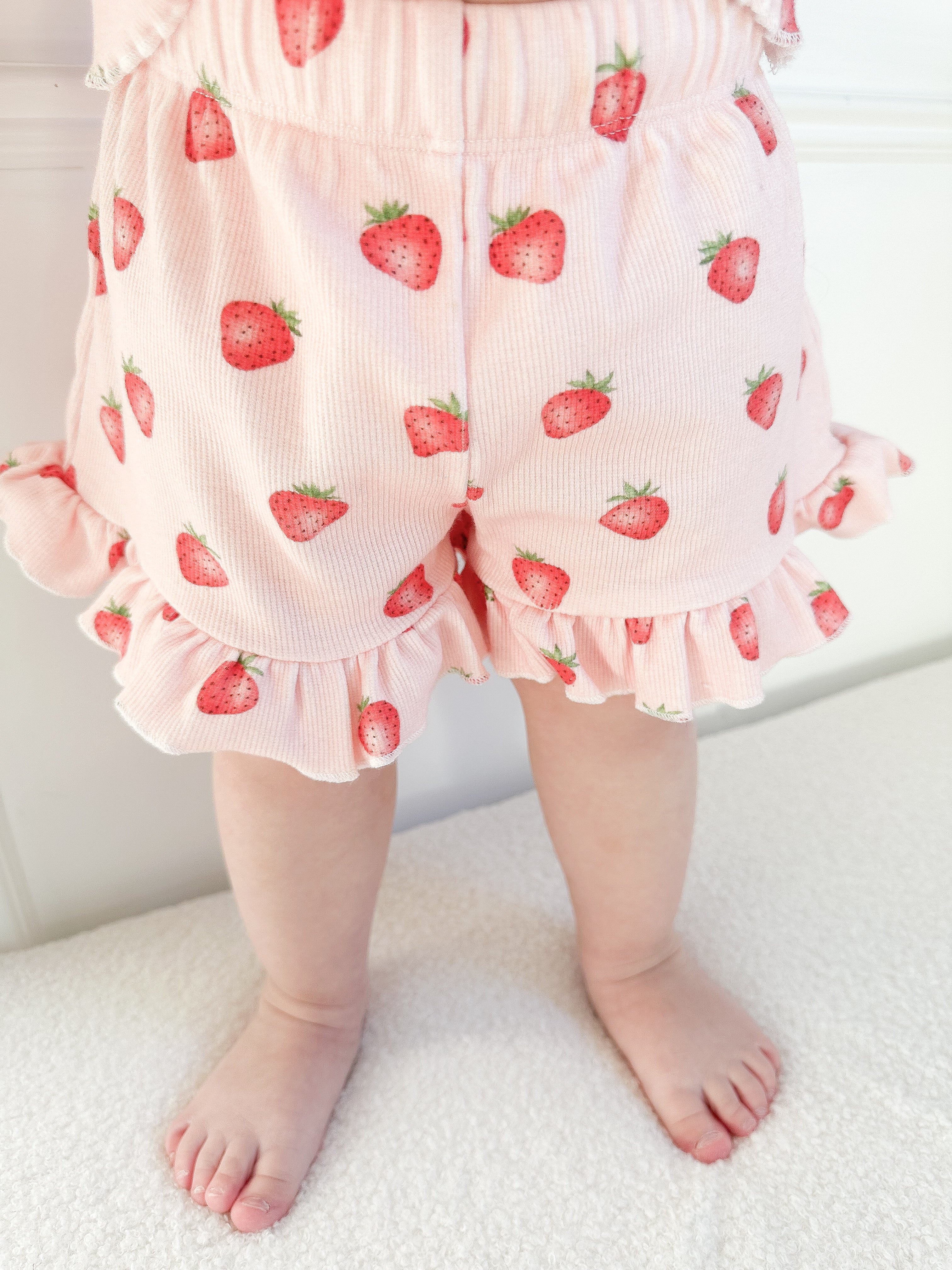 Baby wearing pink ruffle shorts with strawberry pattern standing on a light-colored surface on a white background. Cropped to only show shorts, bare feet.
