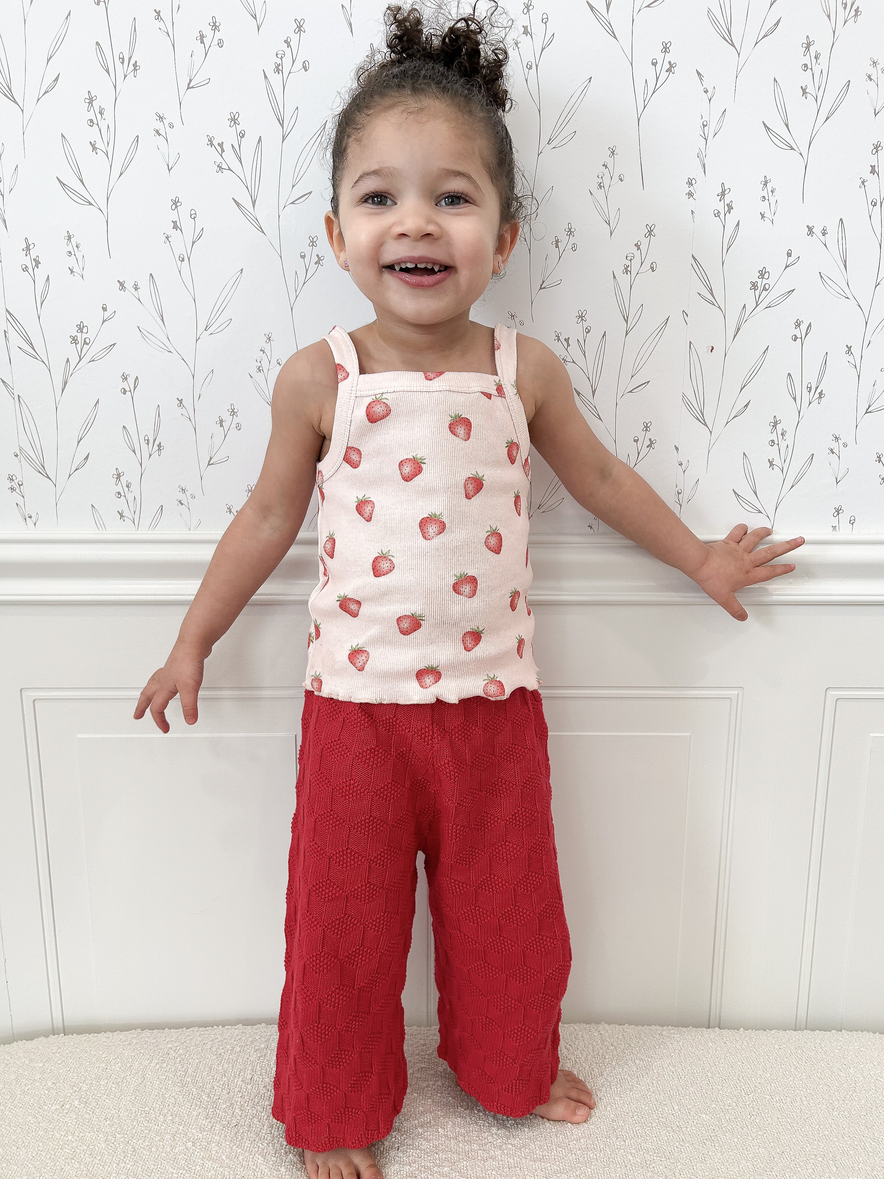 Child wearing a strawberry-patterned tank top and dark pink pants standing against a white molding with subtle floral design above. Child has hair in bun and is smiling and gently touching molding.