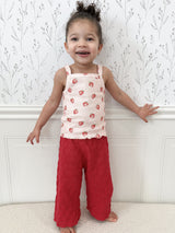 Child wearing a strawberry-patterned tank top and dark pink pants standing against a white molding with subtle floral design above. Child has hair in bun and is smiling and gently touching molding.