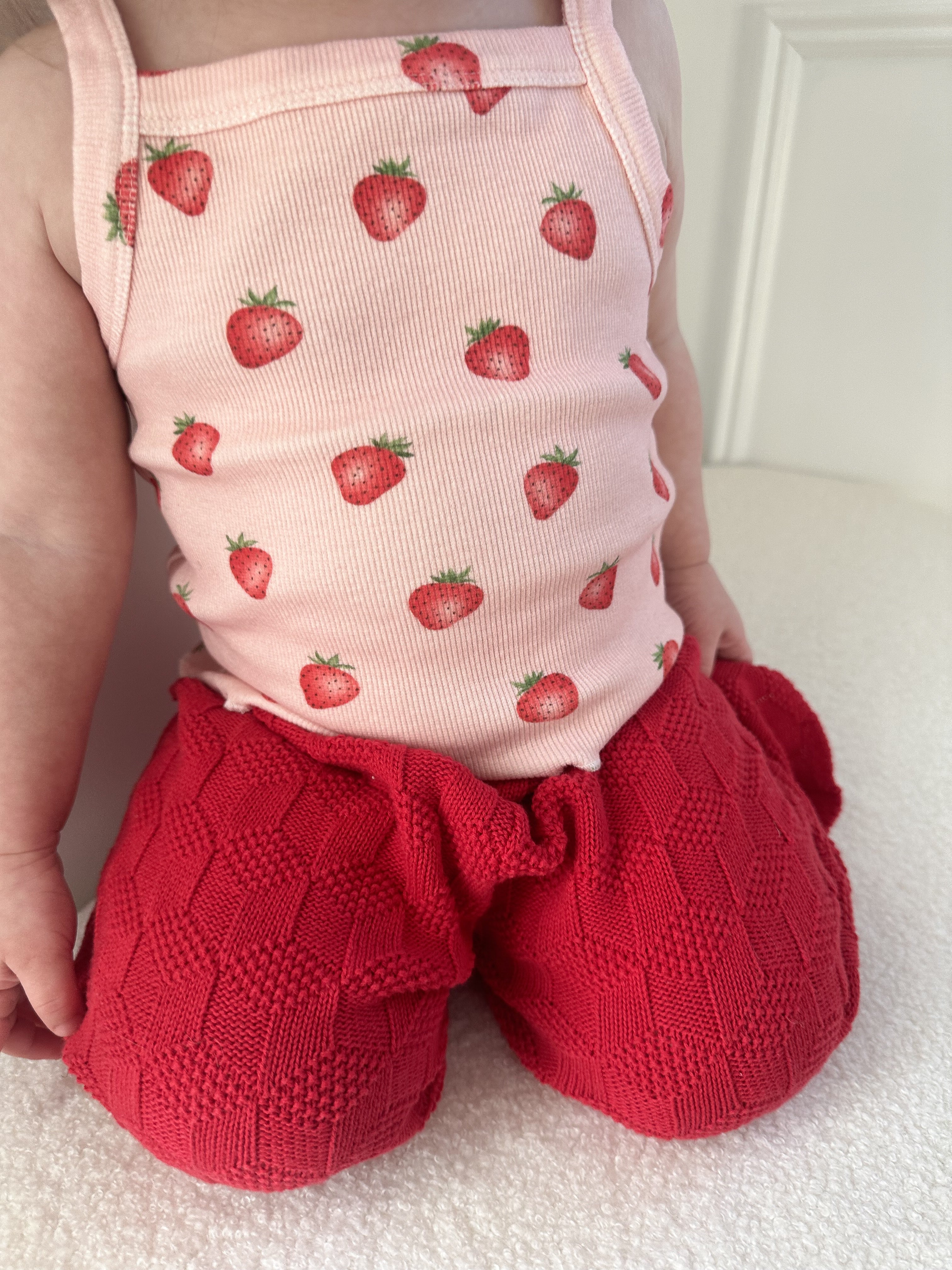 Child wearing a pink sleeveless top with strawberry pattern and red knitted pants against a white background kneeling on a light-colored surface. Cropped to only show outfit.