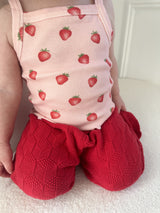 Child wearing a pink sleeveless top with strawberry pattern and red knitted pants against a white background kneeling on a light-colored surface. Cropped to only show outfit.
