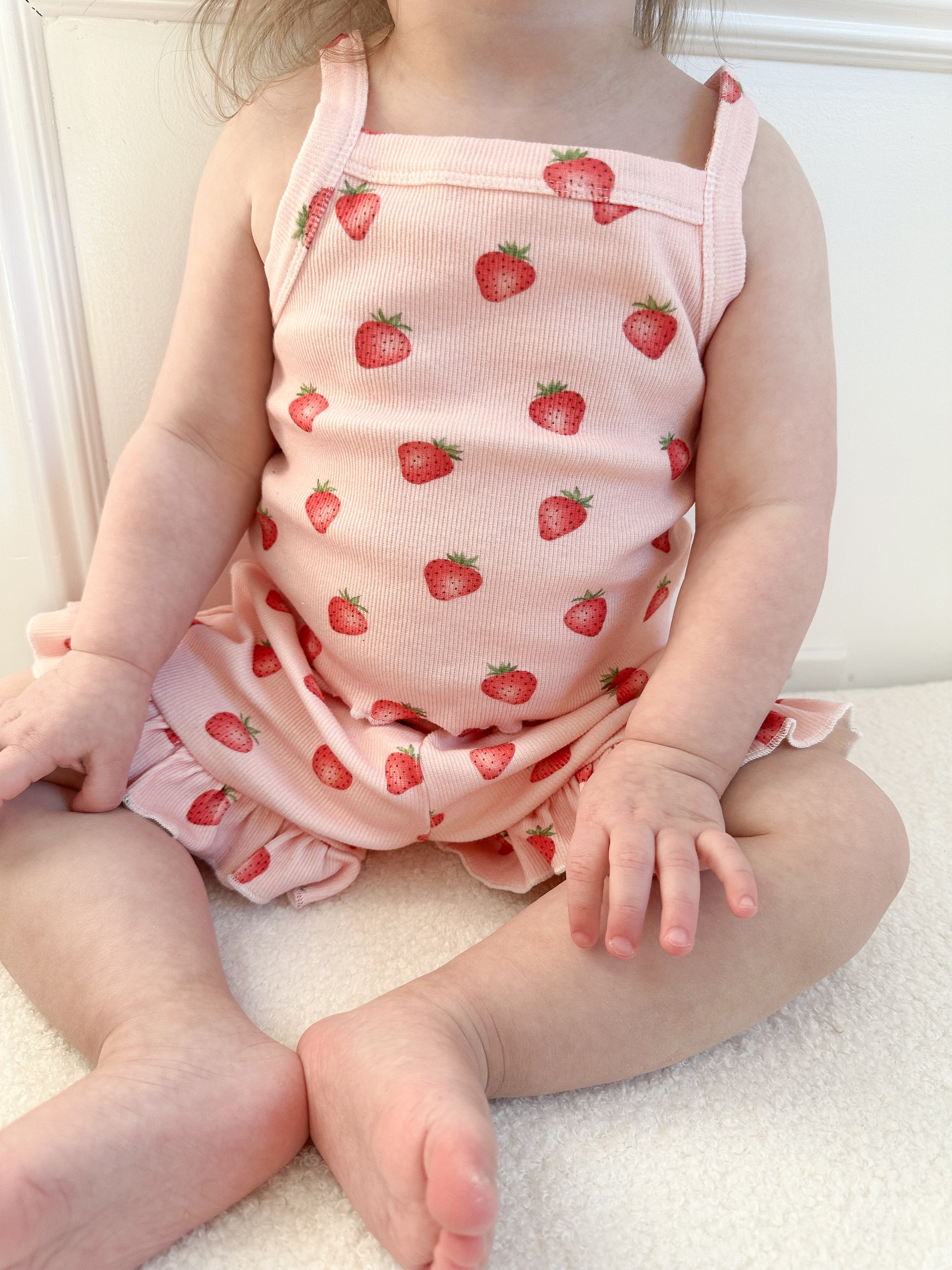 Child wearing a pink ribbed tank top with strawberry pattern and matching ruffle shots sitting on a white surface. Cropped to only show outfit.