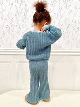 Toddler wearing a blue checkered outfit standing on a white sherpa ottoman facing away with a white paneled wall and wallpaper with decorative flowers in the background.