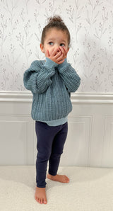 Child wearing a blue checkered sweater and navy leggings standing against a white wall with subtle patterns. Child has hands covering mouth.