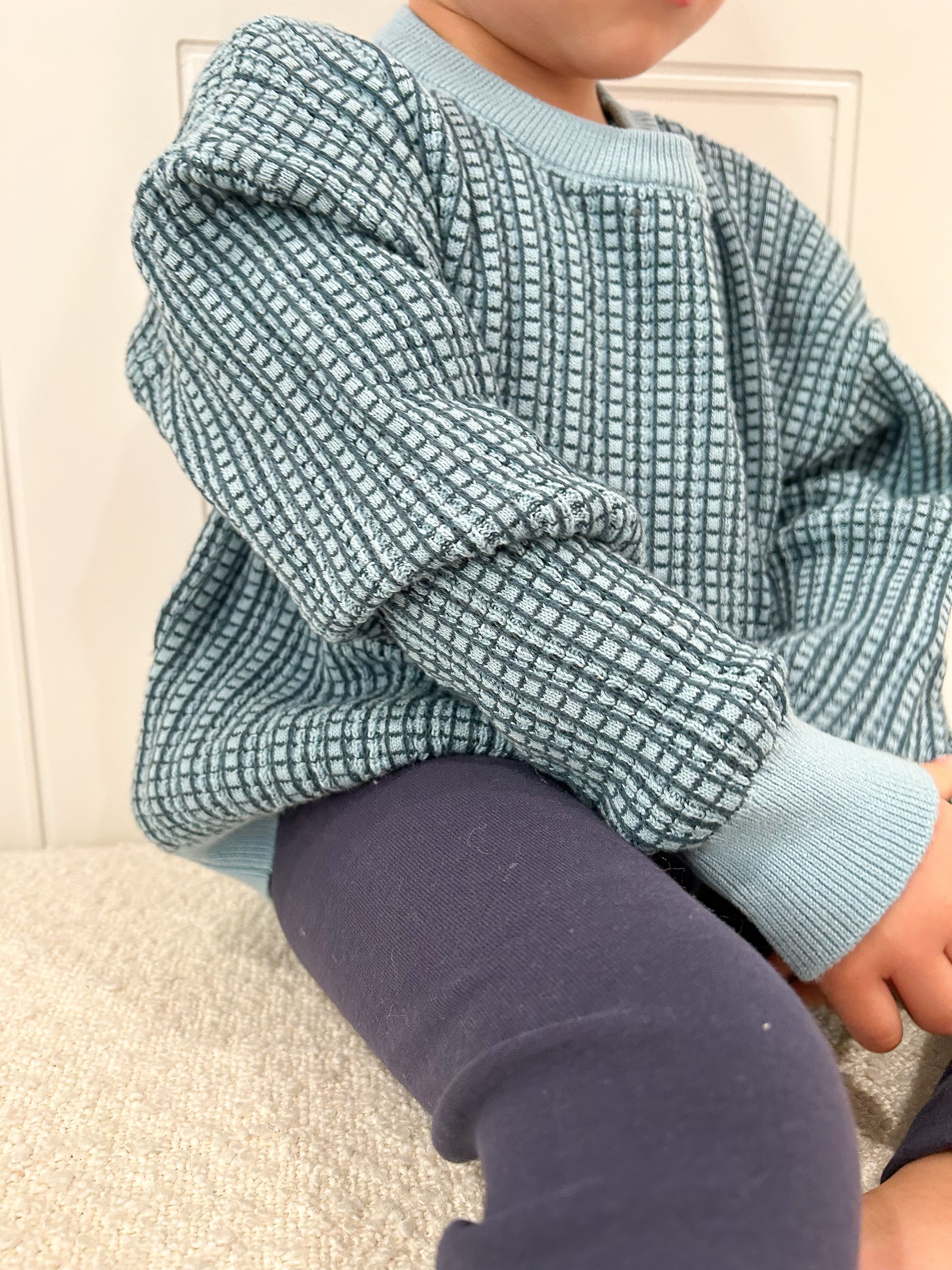 Child wearing a blue and navy checkered shirt with rolled-up sleeves, sitting on a beige couch. Cropped to only show outfit.