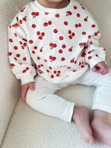 Child wearing a white sweatshirt with red cranberry pattern and white leggings sitting on a beige surface. Cropped to only show outfit.