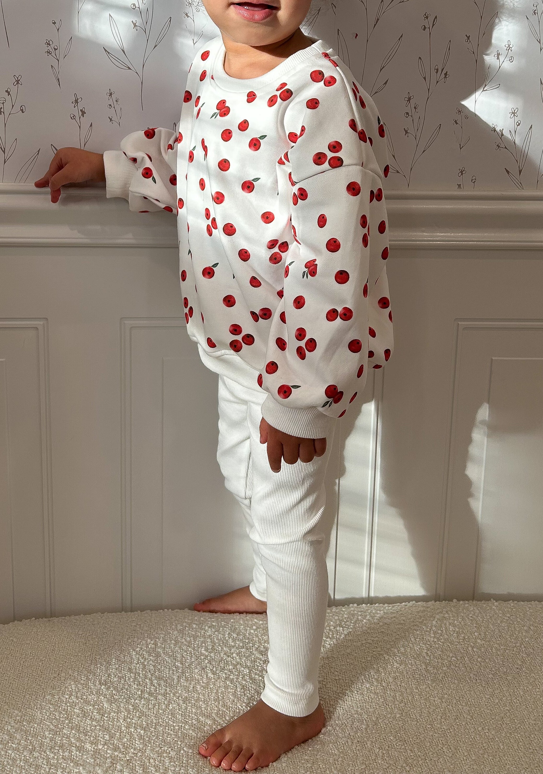 Child wearing a white sweatshirt with cranberries and white leggings standing against floral pattered wall with molding. Cropped to only show outfit.