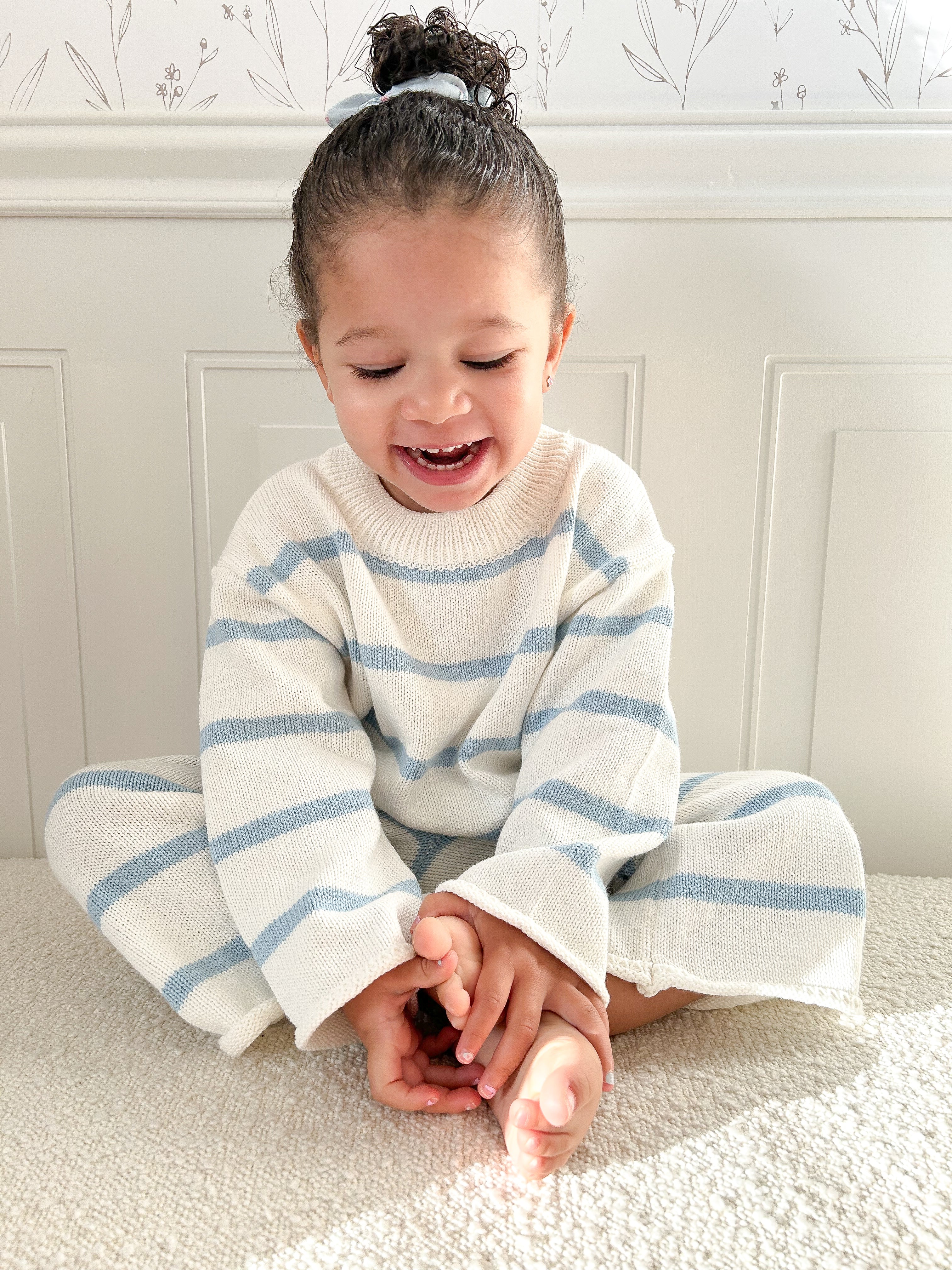 Child in a bright room wearing a white and blue striped outfit sitting on a beige textured surface against white molding and white wall with floral pattern. Child has a bun, is smiling looking down and grabbing feet. 