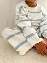 Child wearing a white outfit with blue stripes sitting on a light-colored surface. Cropped to only show outfit.