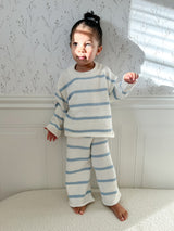 Child in a bright room wearing a white long-sleeve set with blue stripes standing in a room with white walls with a floral pattern and white molding. Child  looking away, has a bun in her hair, light shining and has serious face.