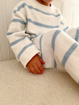 Child wearing a white sweater with blue stripes and matching pants sitting on a beige surface. Cropped to only show outfit.