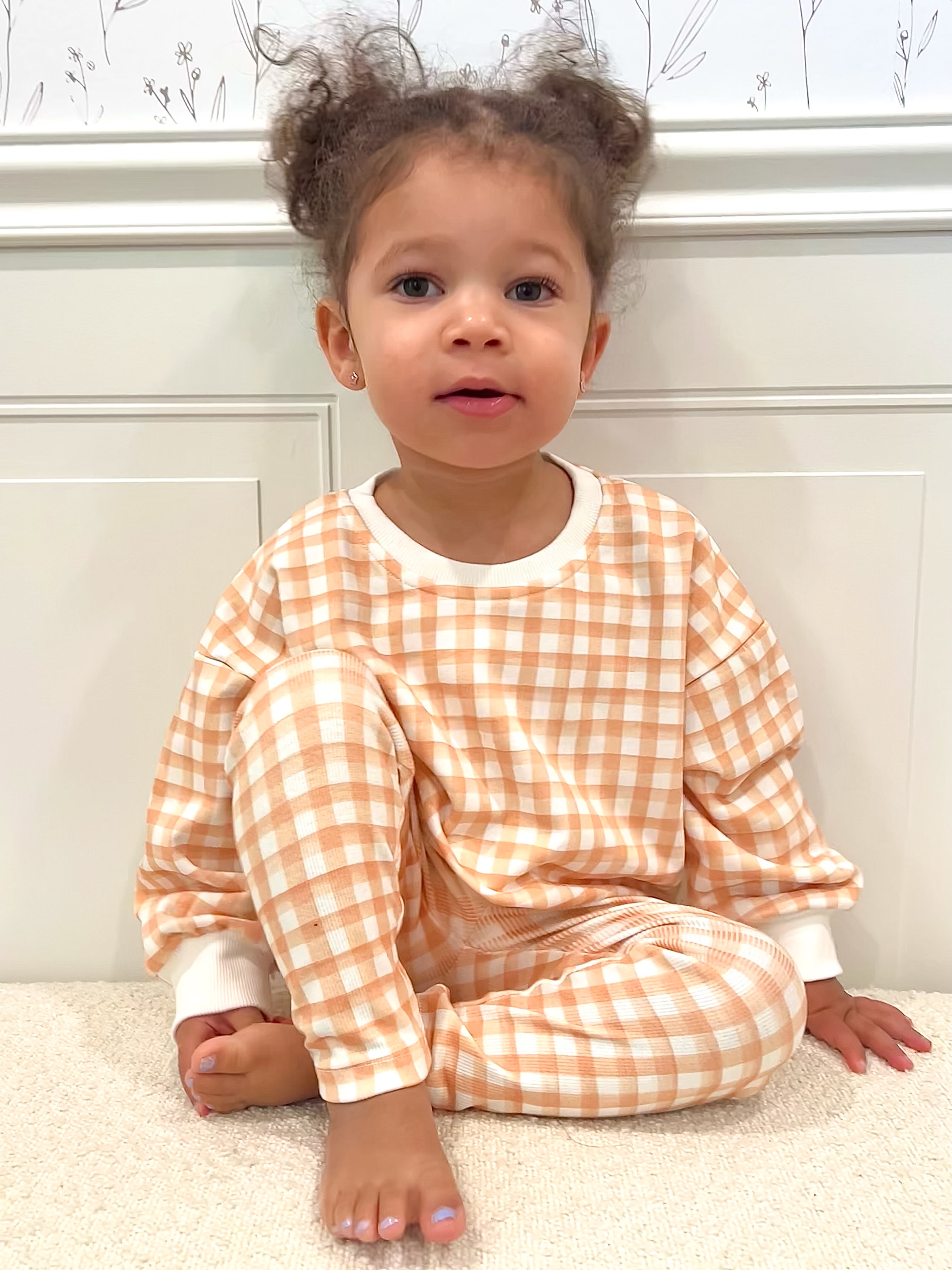 Toddler wearing a checkered outfit sitting on a sherpa ottoman with a neutral background.