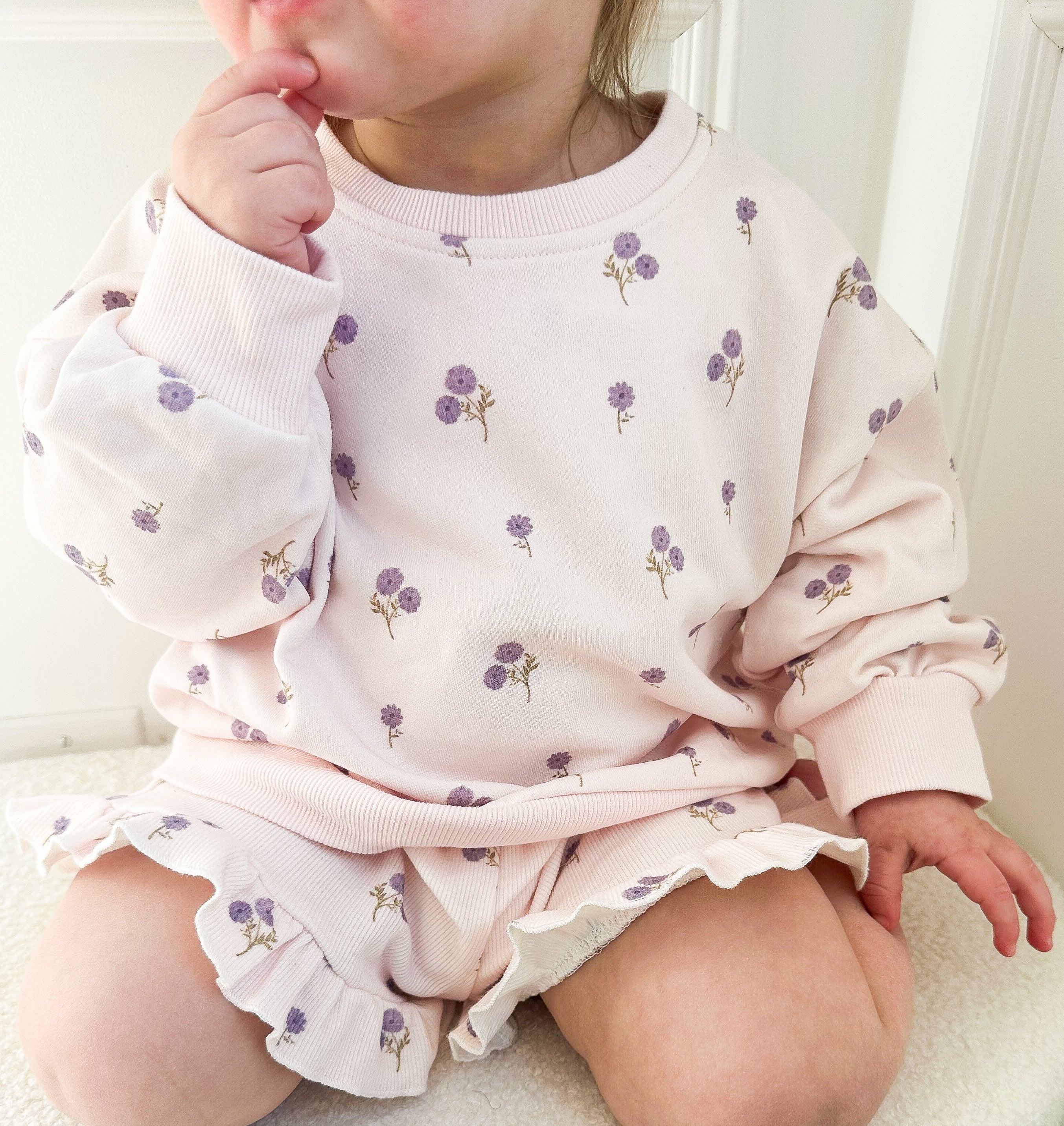 Child wearing a light purple floral outfit sitting on a light beige surface.
