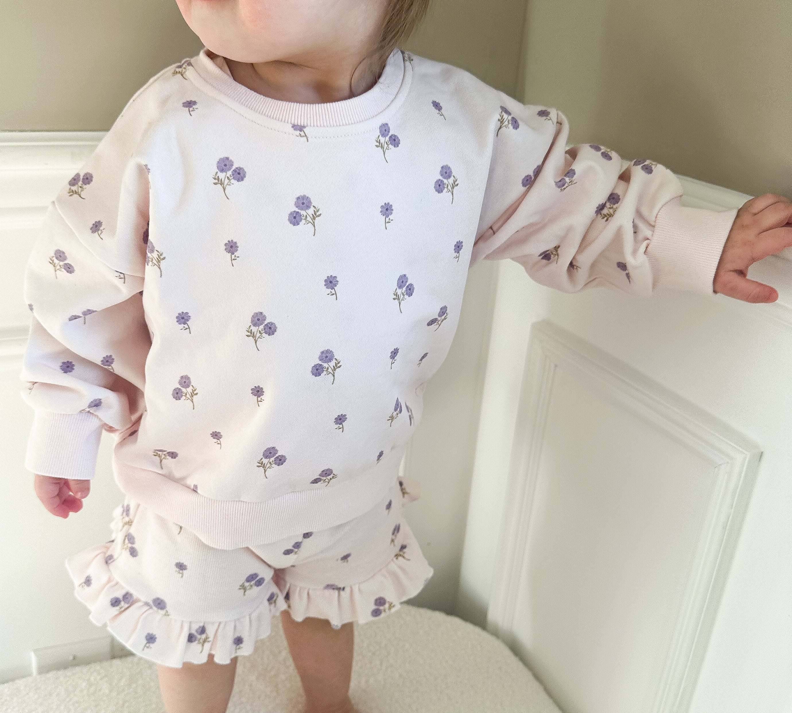 Child wearing a light purple outfit with purple floral patterns in a minimal indoor setting.