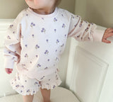 Child wearing a light purple outfit with purple floral patterns in a minimal indoor setting.