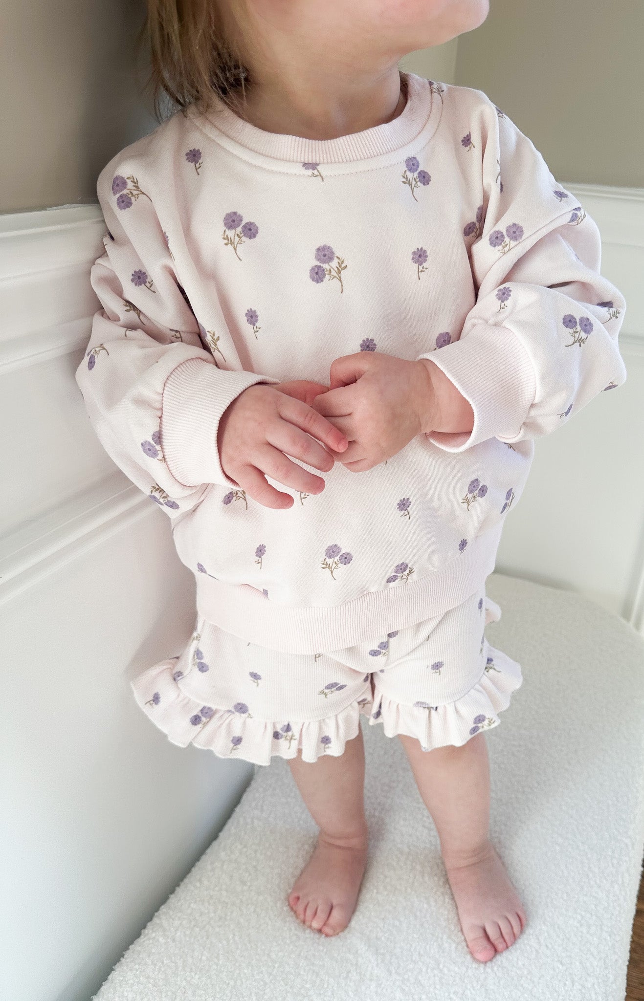 Child wearing a light purple floral long sleeve and ruffle shorts outfit set standing on light-colored surface.