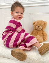 Toddler in pink and purple striped outfit with teddy bear sitting on light-colored surface against white background. Toddler is turned away with her face turned toward camera smiling.