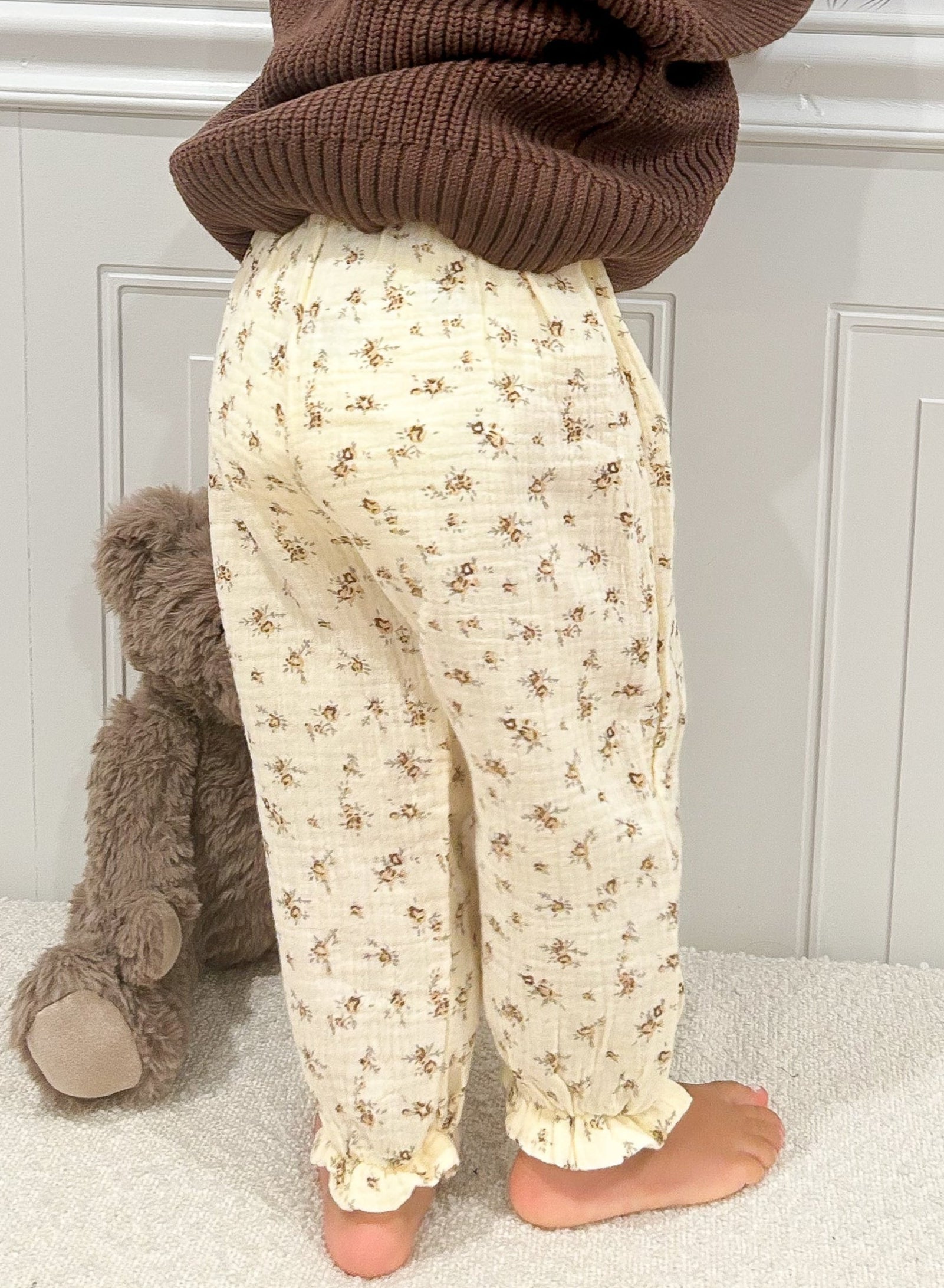 Beige pants with brown floral pattern on toddler, there is half a teddy bear in image against white wall 