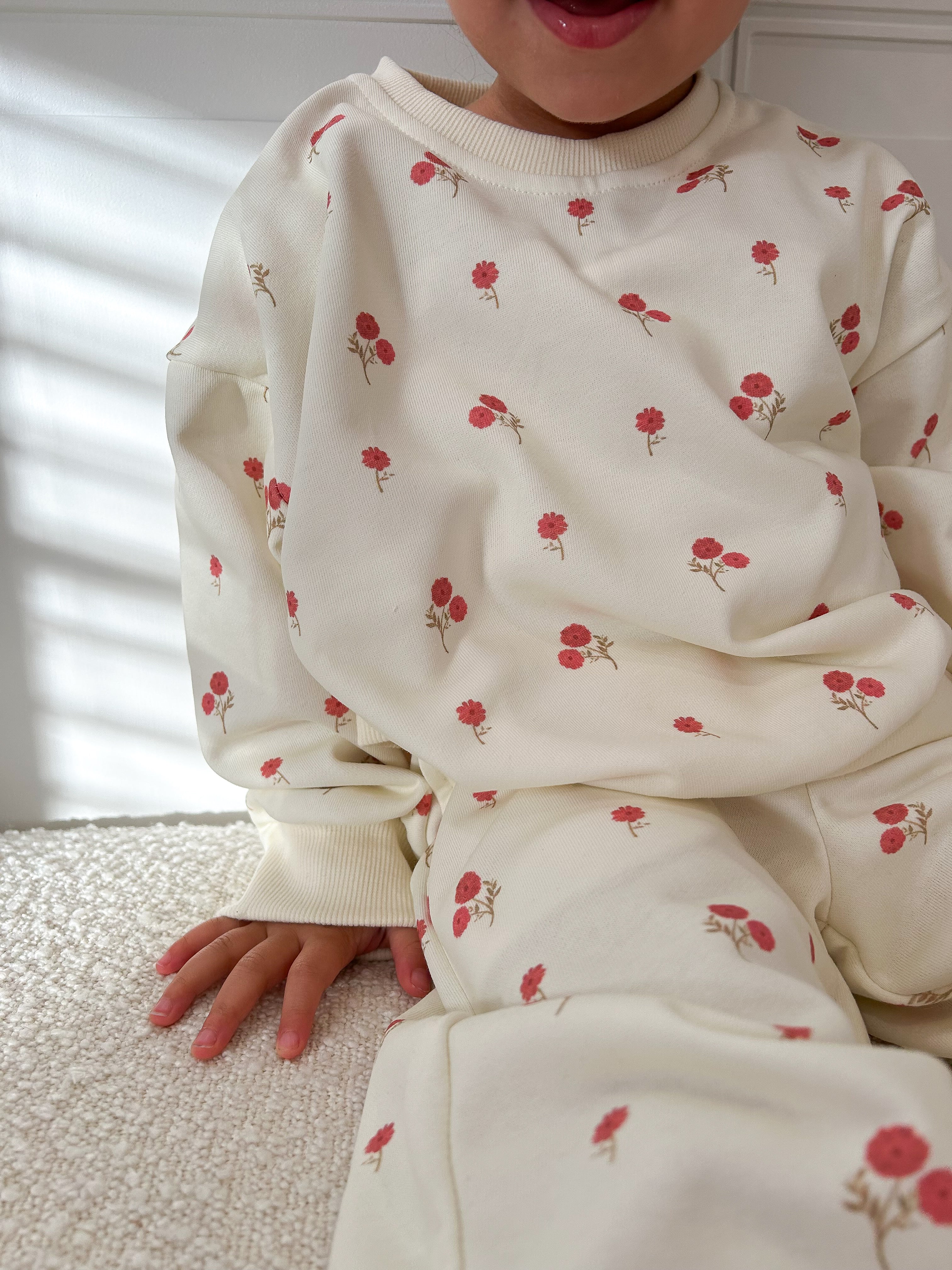 Child wearing a cream-colored outfit with red floral patterns sitting on a textured surface. Cropped to only show outfit.