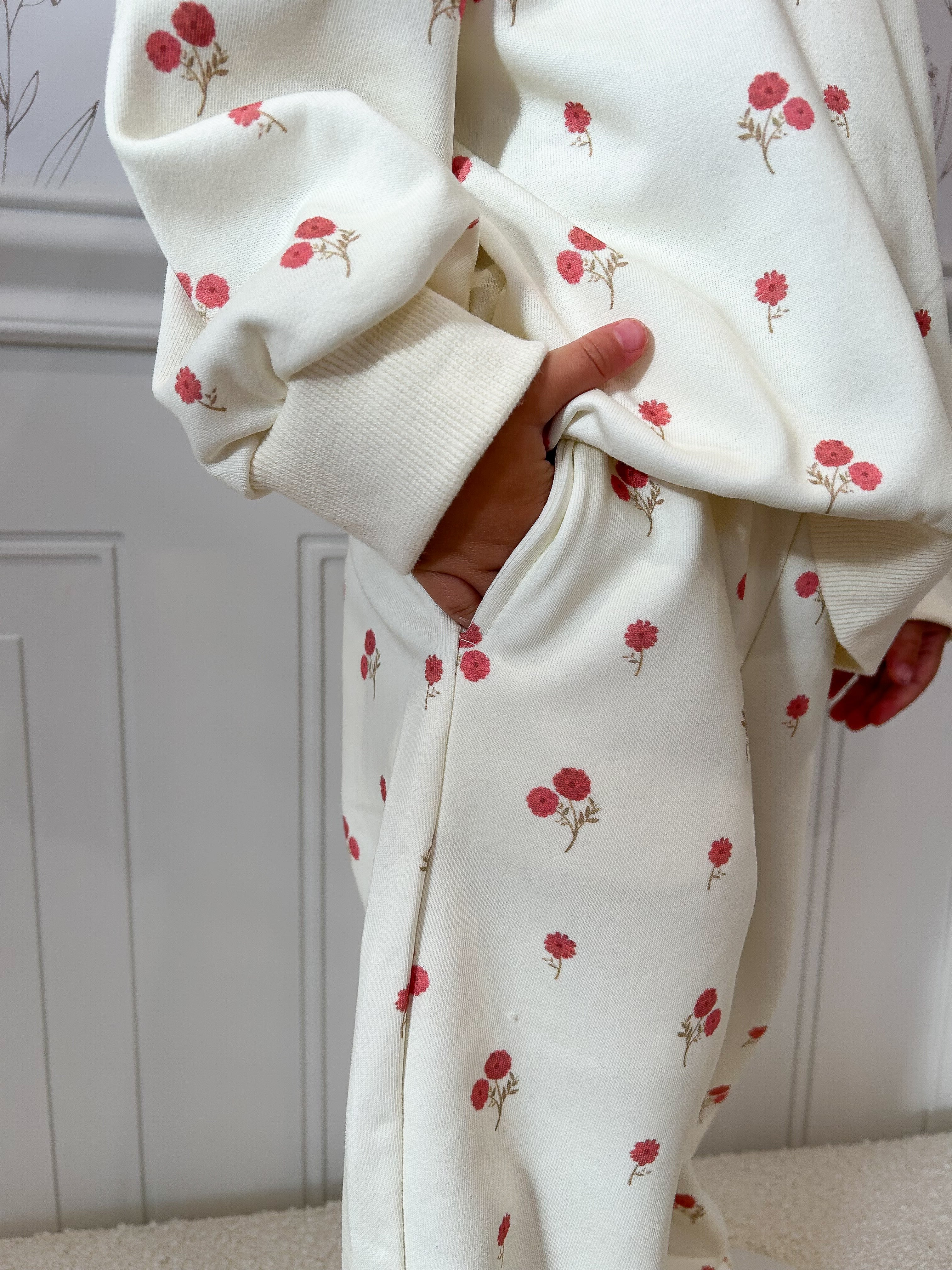 White baby sweat set with red floral patterns worn by a child, against a neutral background. Childs hand is in pocket.