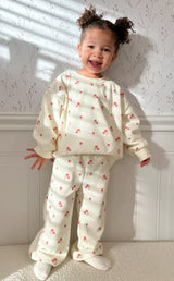 Child wearing a cream-colored outfit with red floral patterns in a room with white walls with flowers and molding standing on a light-colored surface. Child is smiling and has two buns in hair.