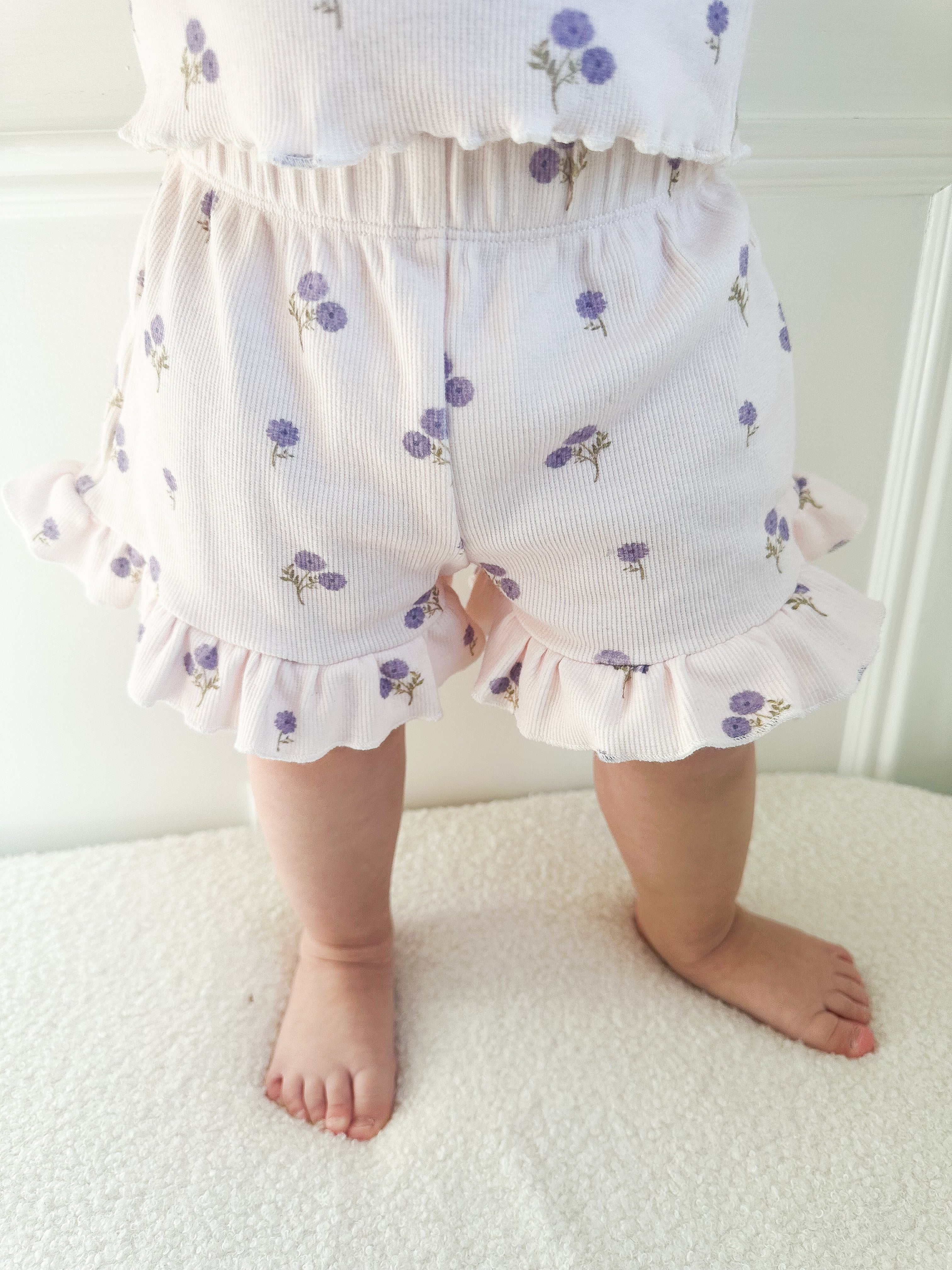 Baby wearing light-purple shorts with purple floral patterns and ruffle trim standing on a light surface.