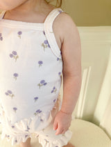 Child wearing a light-purple tank with floral patterns with matching shorts with ruffle trim standing on a light-colored surface.